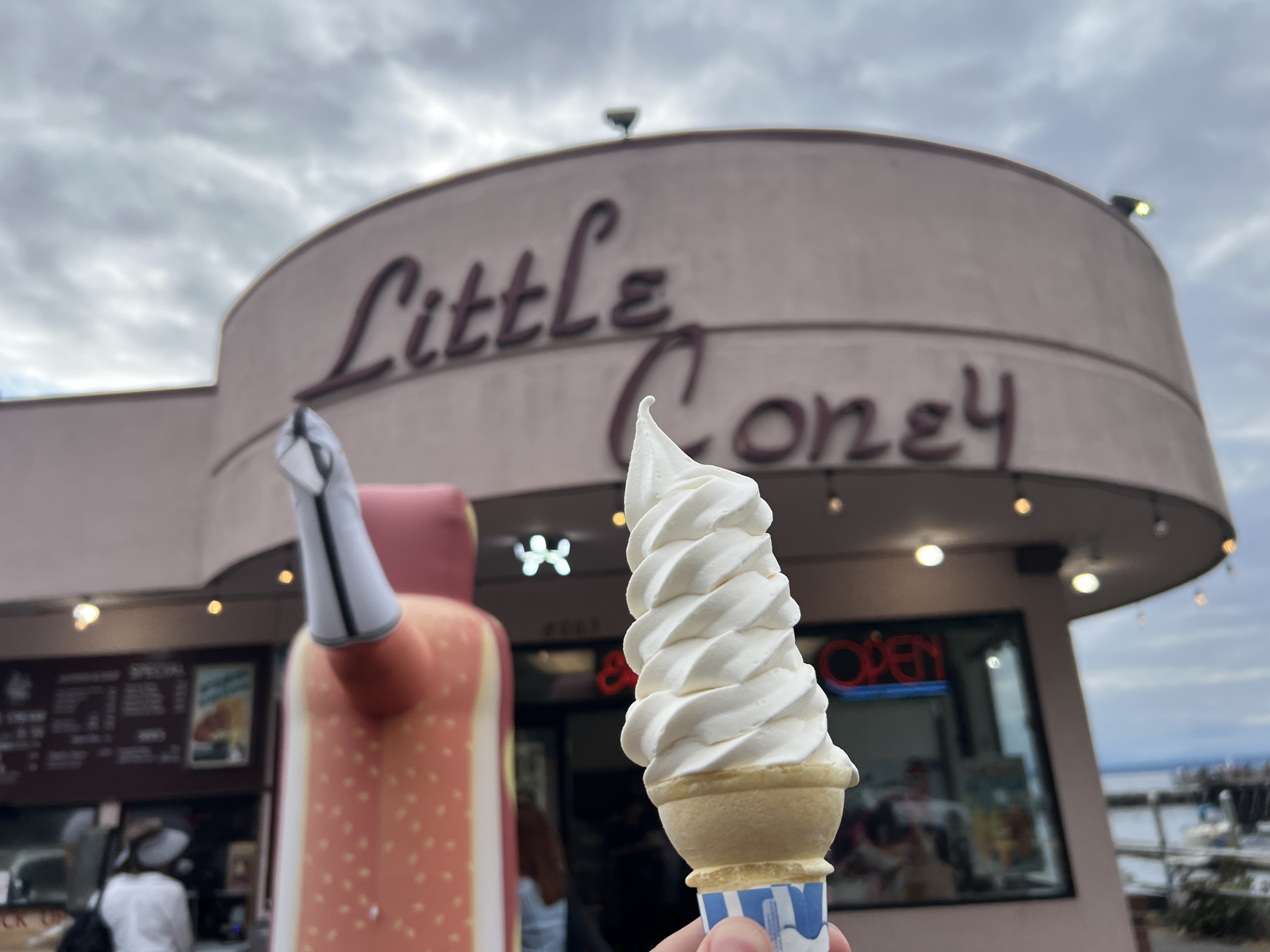 A pure white swirl of vanilla soft serve in a cone. In the background is a sign in italic text that says "Little Coney".