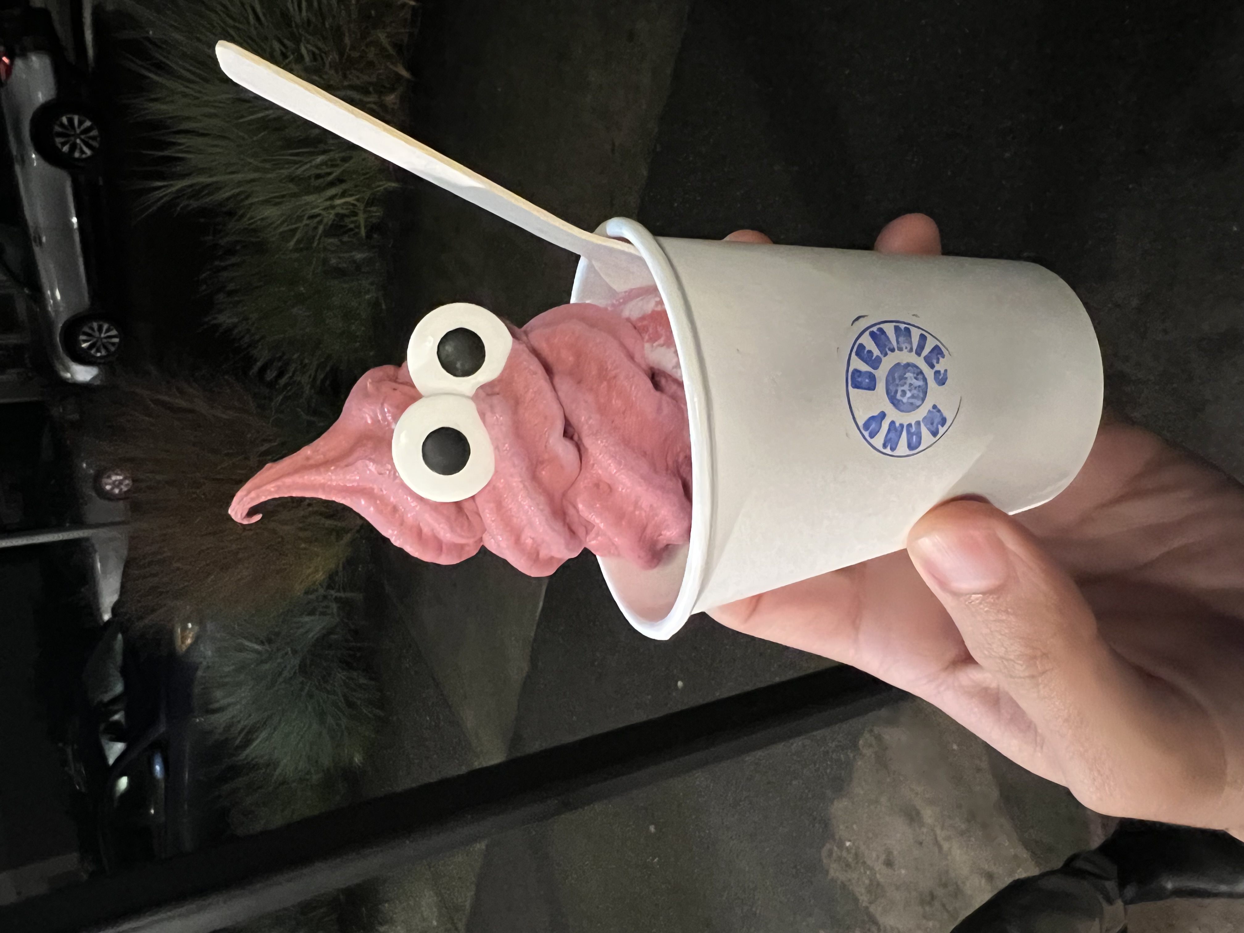 A pink swirl of soft serve with two googly eyes. The soft serve is perched in a paper cup with the Many Bennies logo stamped on the front.